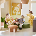 A 1:12 scale Sweetwood Cottage Dollhouse living room scene featuring four wooden figures, including a parent holding a baby on a sofa, a seated child, and a standing child.