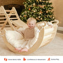 A smiling toddler sits inside a wooden curved rocker fitted with a Thick Padded Play Cushion, with a wooden climbing triangle and a decorated Christmas tree in the background.