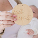 A person with pink and white swirled nails holds a round wooden sign reading "Tiny Land Hello World," while a baby dressed in white and Tiny Land® Montessori Toys for Newborns (0–3 Months) appear in the background.