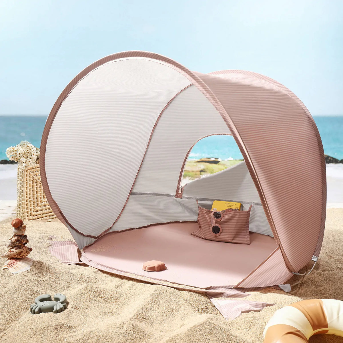 Blush pink Kids Beach Tent & Sand Toy Set with a mesh window and interior storage pocket, surrounded by a sand toy set on a beach.