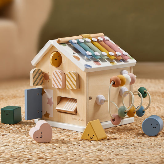 The Montessori Cottage Activity Cube – Morandi is a wooden house-shaped toy featuring a xylophone roof, bead maze, and interactive doors, surrounded by small geometric blocks with smiley faces to help develop fine motor skills.