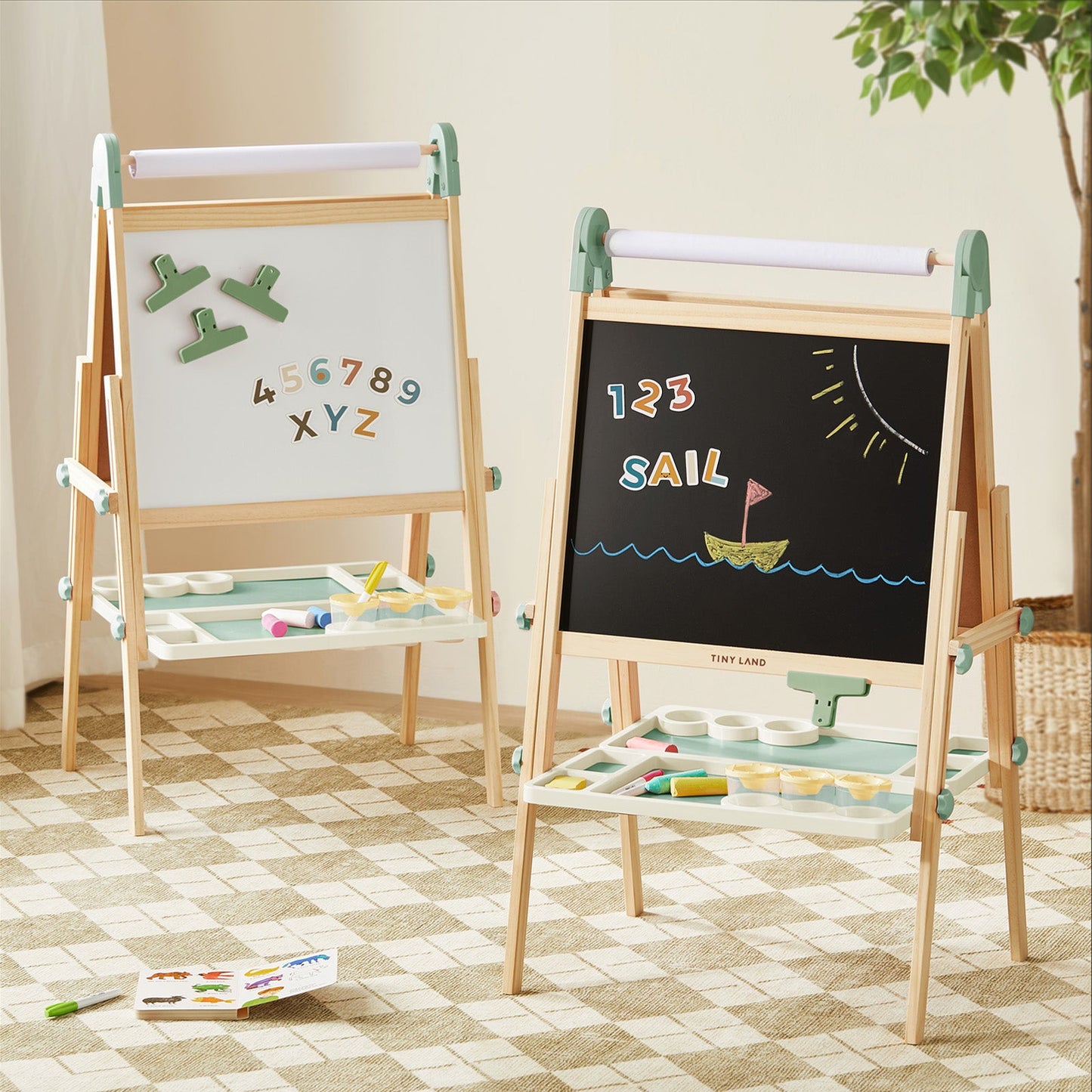 Two Tiny Land® Magnitales Easels for Kids – Green hold art supplies on built-in trays. One magnetic easel displays numbers and letters; the other, a blackboard, shows "123 SAIL" with boat, sun, and waves. Both stand on a checkered rug.