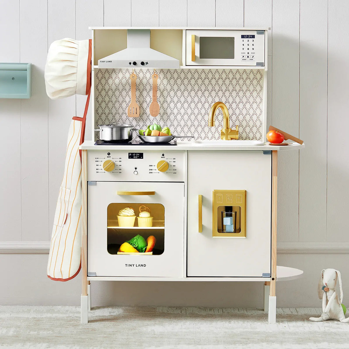 The PopJoy Play Kitchen with Lights & Sounds is a white play kitchen with gold accents, featuring a stove, microwave, sink, storage compartments with pretend accessories, and a chef hat and apron hanging on the side.