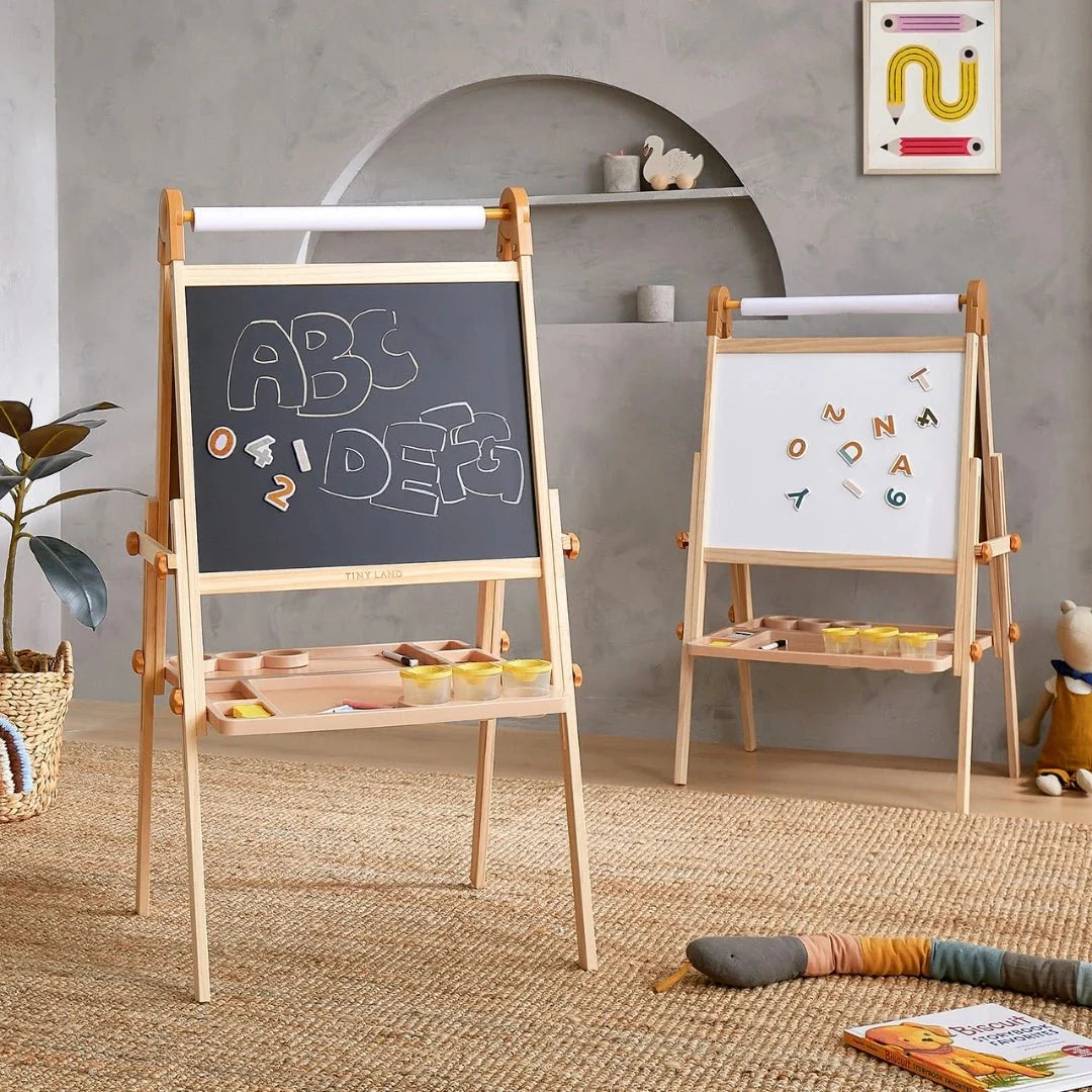 Two Tiny Land® Magnitales Easels for Kids stand in a cozy playroom: one has a chalkboard with “ABC IDE?” written on it, and the other is a magnetic art easel showing scattered letters. Toys and art supplies are neatly organized around the room.