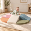 A baby crawls on a PlayNest Soft Climbing Blocks & Ball Pit Set – Morandi, a modular set of pastel foam steps, a ramp, and a wedge in an indoor playground.