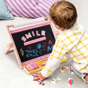 A child plays with a 3-in-1 Adjustable Tabletop Art Easel featuring a blackboard decorated with magnetic letters spelling SMILE, hand-drawn chalk art, and a storage tray holding an eraser and chalk.