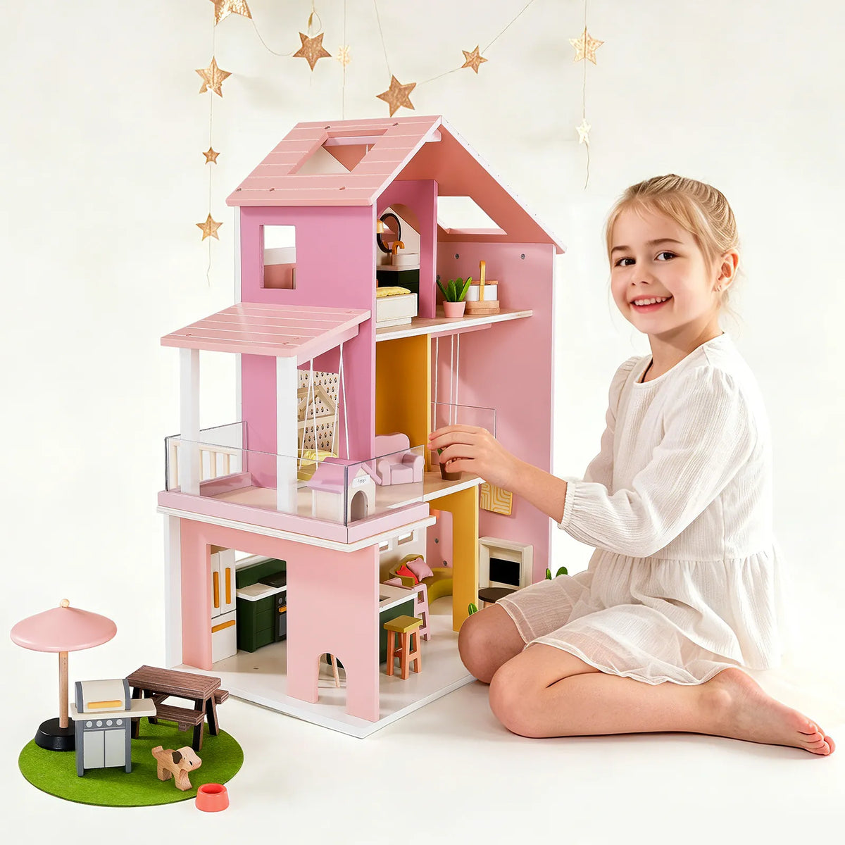 A young girl smiles while playing with a pink and white Sweetwood Cottage Dollhouse, featuring 1:12 scale furniture and a small dog figure arranged on a green mat nearby.