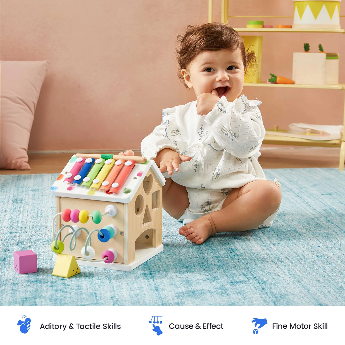 A baby sits on a blue rug playing with a Montessori Cottage Activity Cube – Rainbow, which features a xylophone, bead maze, and shape sorter to help develop fine motor skills.
