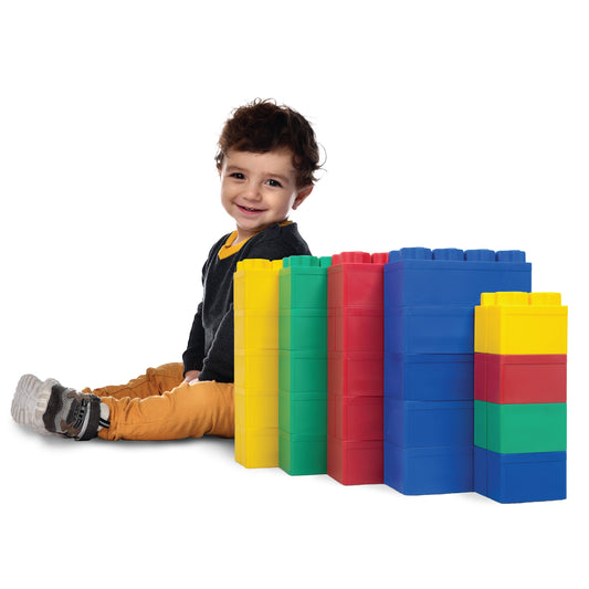 A young child sits on the floor smiling next to several large, colorful Jumbo Building Blocks 24 PC - Beginner Set in yellow, blue, green, and red against a white background.