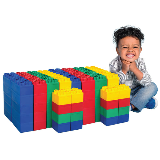 A smiling young child sits beside colorful red, blue, green, and yellow Jumbo Building Blocks 96 PC - Standard Set, stacked in rows on the floor. The excited child looks happy next to the jumbo blocks.