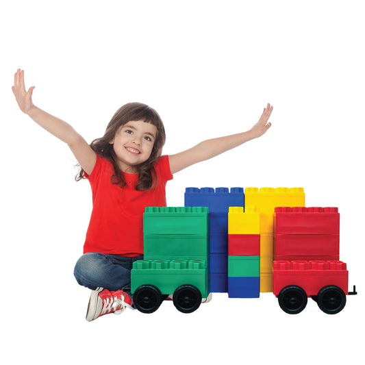 A smiling young girl in a red shirt sits with arms raised behind the Jumbo Building Blocks 24 PC with 2 Wheel Kits - Big City Set and block vehicles on a white background.
