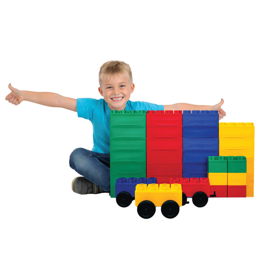 A smiling child in a blue shirt sits on the floor, arms outstretched behind a set of Jumbo Building Blocks 36 PC with 3 Wheel Kits - Train Set, including colorful giant blocks and a toy car built from the same pieces.