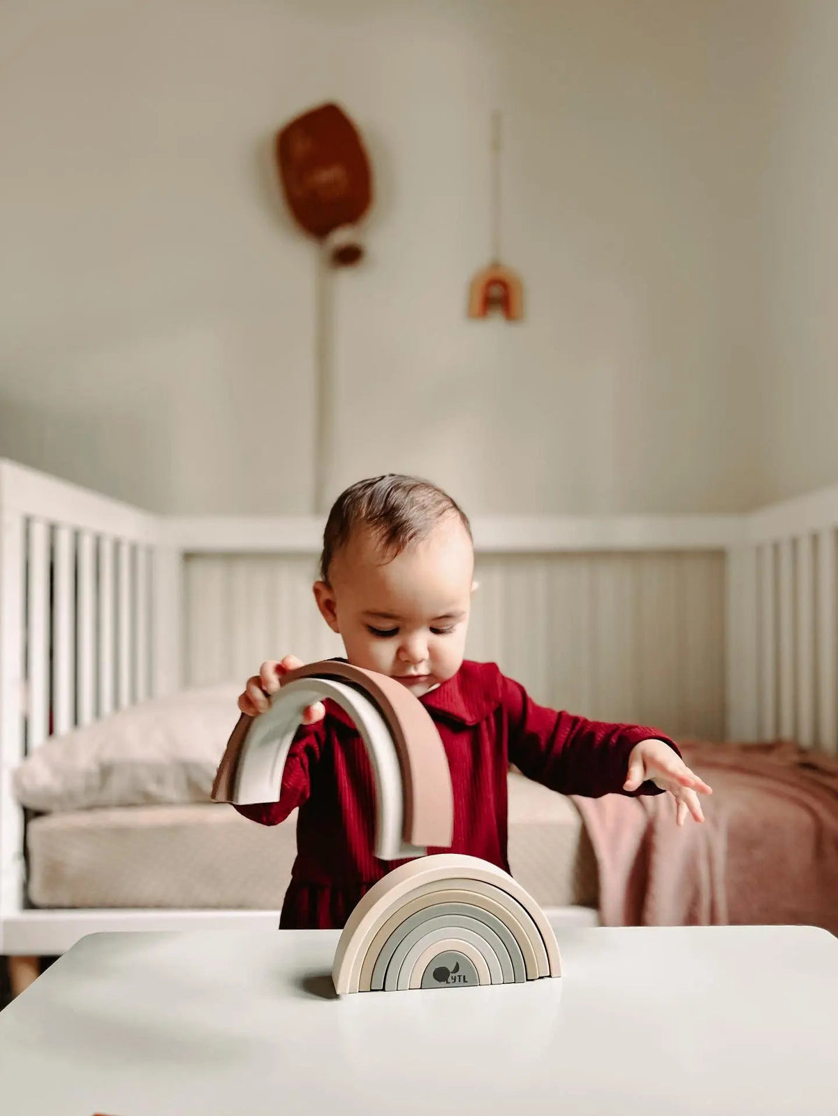 A toddler in a maroon dress stacks a 10 pc Silicone Rainbow Stacking Toy on a white table.