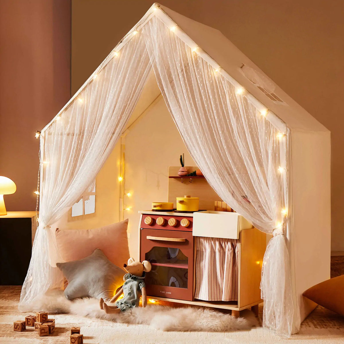A Large Cozy Cabin Play Tent with Star Lights featuring sheer curtains, a toy kitchen, a stuffed mouse, and alphabet blocks on the floor.