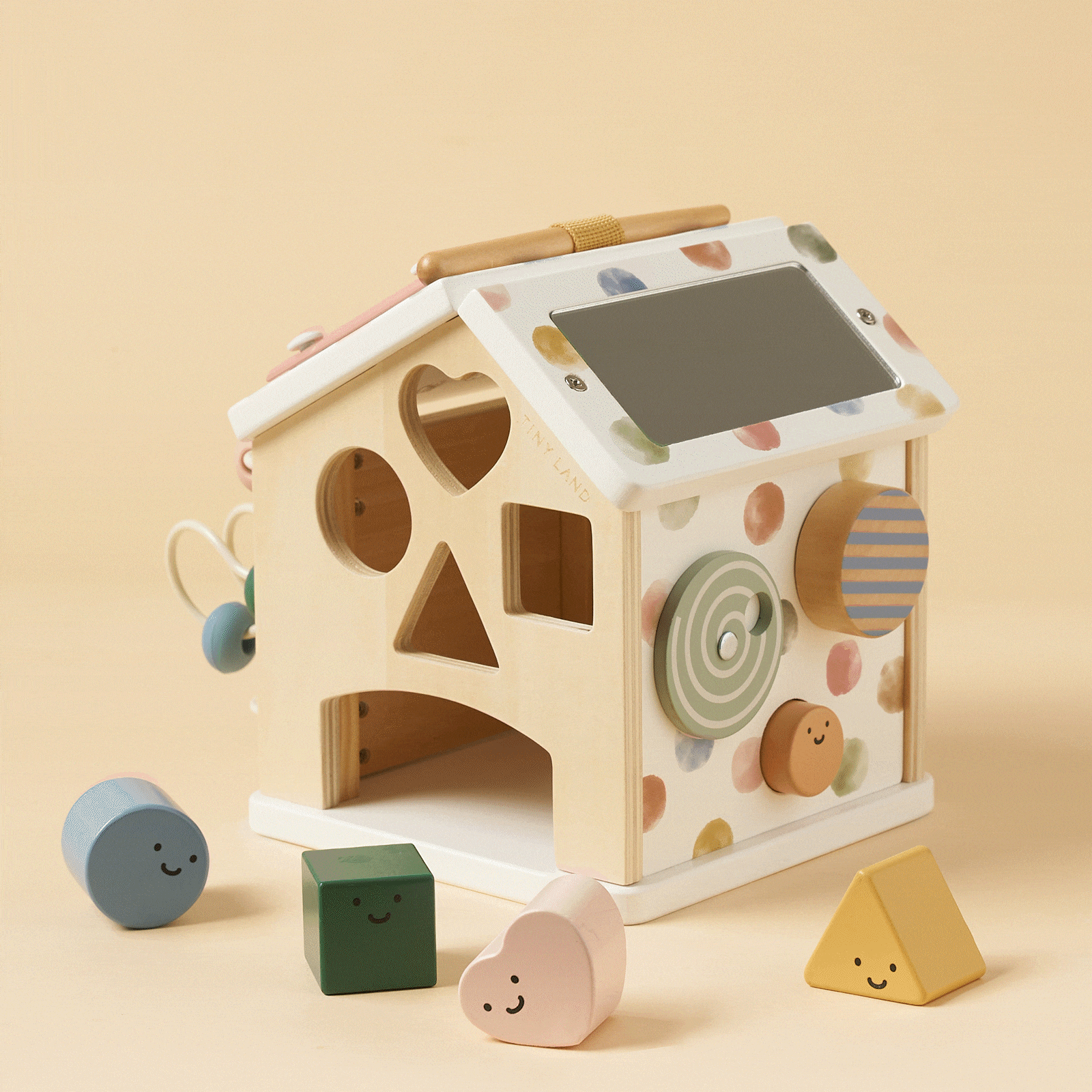 The Montessori Cottage Activity Cube – Morandi is a wooden house-shaped activity cube with shape-sorting cutouts, a mirror, and spinning gears, surrounded by four small colorful blocks with smiling faces to help develop fine motor skills.