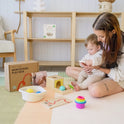 A woman and a baby play on a mat with Tiny Land® Montessori Toys for Newborns (7–9 Months), including stacking rings, a shape sorter, sensory ball, book, Montessori Play Box, and Object Permanence Box. She helps the baby explore the toys.