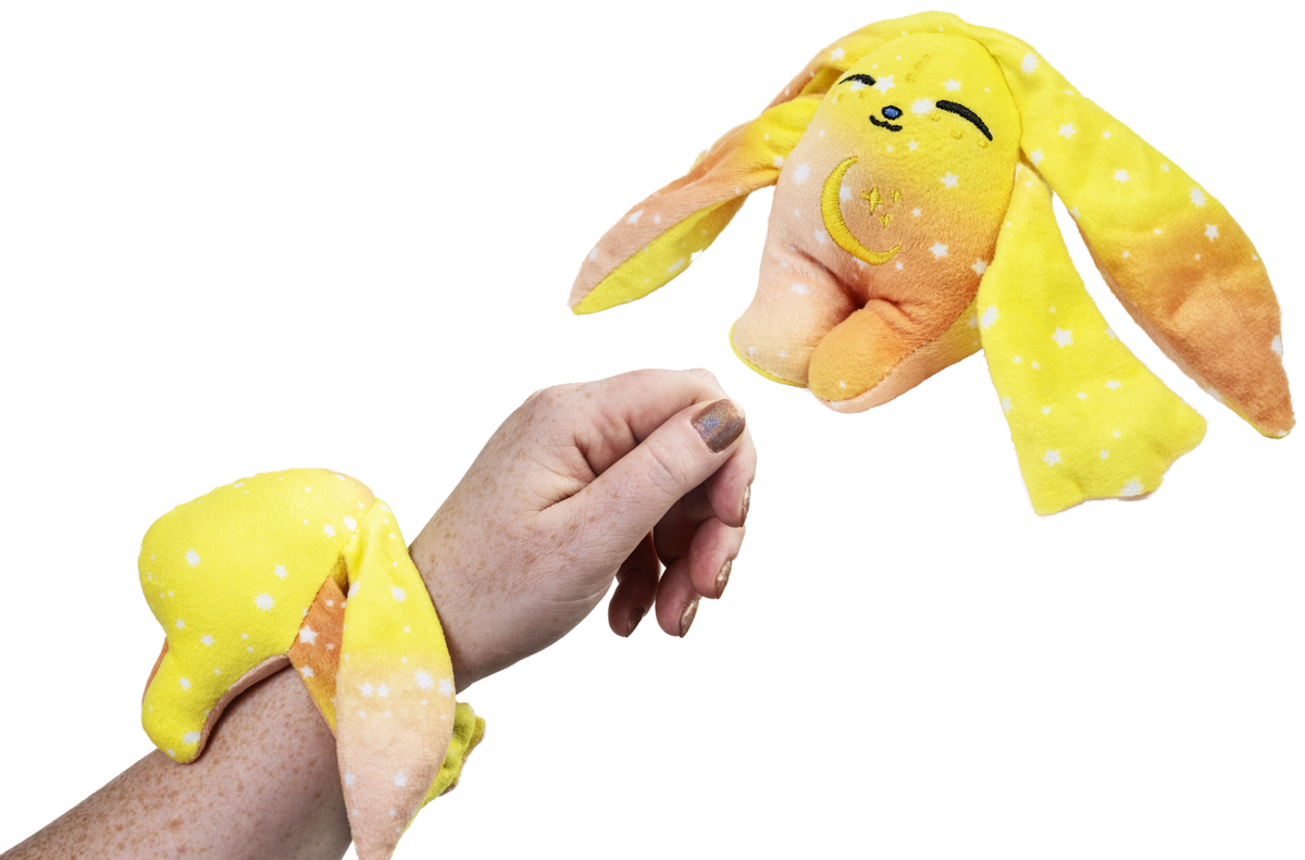 A hand wears a yellow, orange, and white Spark Mini Wrist Hugger – Magnetic Sensory Plush with long ears, while a matching second plush hangs nearby.
