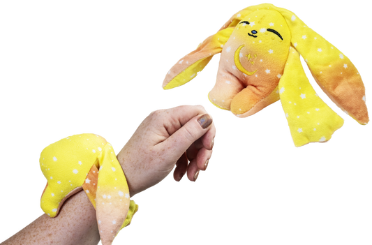 A hand wears a yellow, orange, and white Spark Mini Wrist Hugger – Magnetic Sensory Plush with long ears, while a matching second plush hangs nearby.