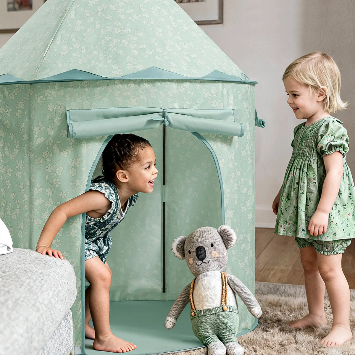 A child peeks out of a hexagonal, light green floral Starry Indoor Play Tent for Toddlers while another child stands nearby with a stuffed koala.