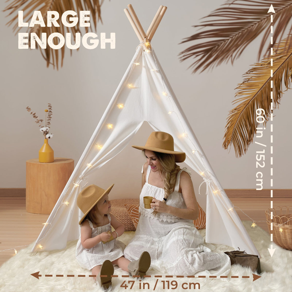 A woman and a young girl in matching white dresses and hats sit inside a Cozy Kids Teepee Tent with Padded Mat, a 60-inch high, 47-inch wide teepee decorated with string lights.