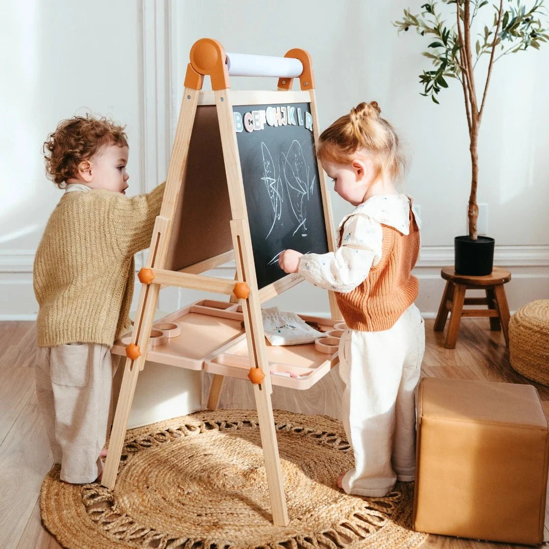Two young children draw on the Tiny Land® Magnitales Easel for Kids, a height-adjustable blackboard, in a cozy, sunlit room featuring light-colored rugs, wicker furniture, and a potted plant in the background.