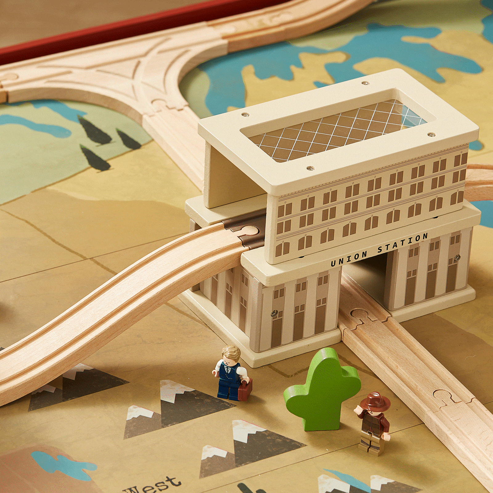 Wooden train tracks from a 1920s Railway Wooden Train Table with Sounds wind through a Union Station building on a vintage city scene map, with two small toy figures standing nearby.