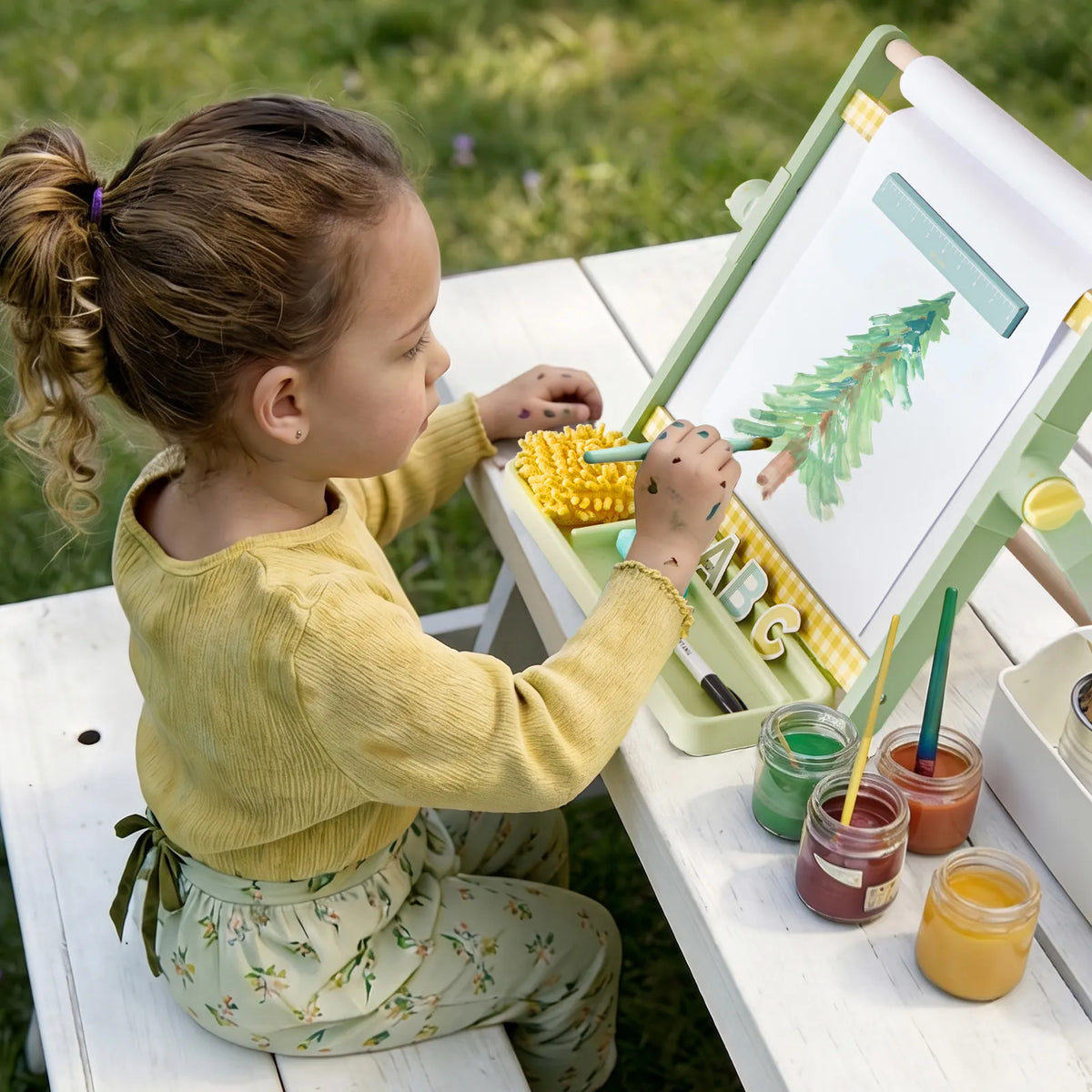 A child sits at a 3-in-1 Adjustable Tabletop Art Easel painting a picture of a pine tree, with jars of colored paint nearby.