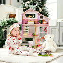 A toddler in patterned pajamas sits on a rug next to a Sweetwood Cottage Dollhouse (48-pc Furniture) and a large white teddy bear.