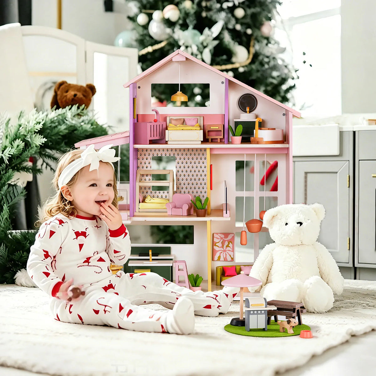 A toddler in patterned pajamas sits on a rug next to a Sweetwood Cottage Dollhouse (48-pc Furniture) and a large white teddy bear.