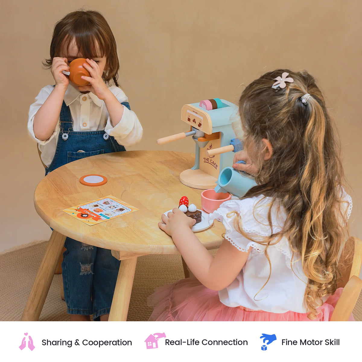 Two young children sit at a small wooden table playing with a Wooden Play Coffee Maker Set, a toy espresso machine, a play menu, and toy pastries.
