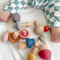 A baby in checkered pants sits on a white surface surrounded by a Mushroom Mix & Match Sorting Toy (24 pc), featuring colorful wooden mushrooms designed to help develop fine motor skills.
