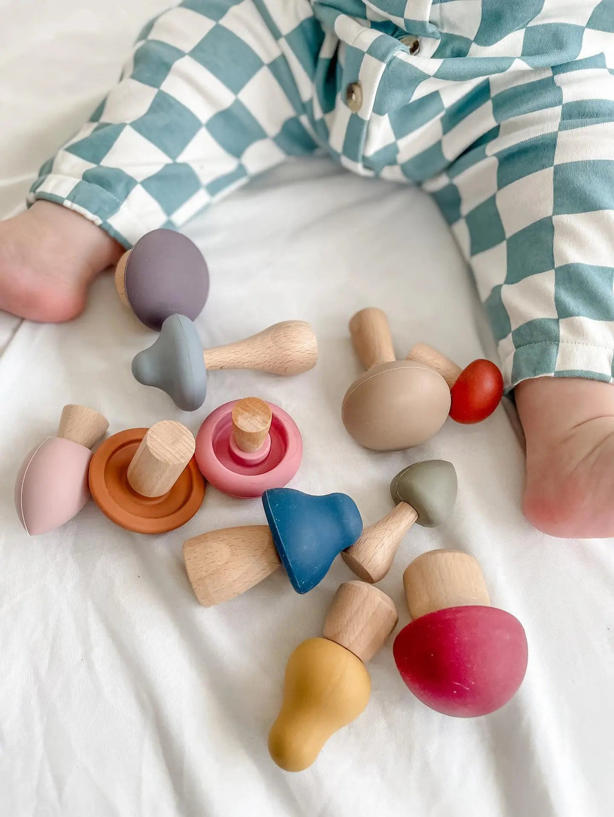 A baby in checkered pants sits on a white surface surrounded by a Mushroom Mix & Match Sorting Toy (24 pc), featuring colorful wooden mushrooms designed to help develop fine motor skills.