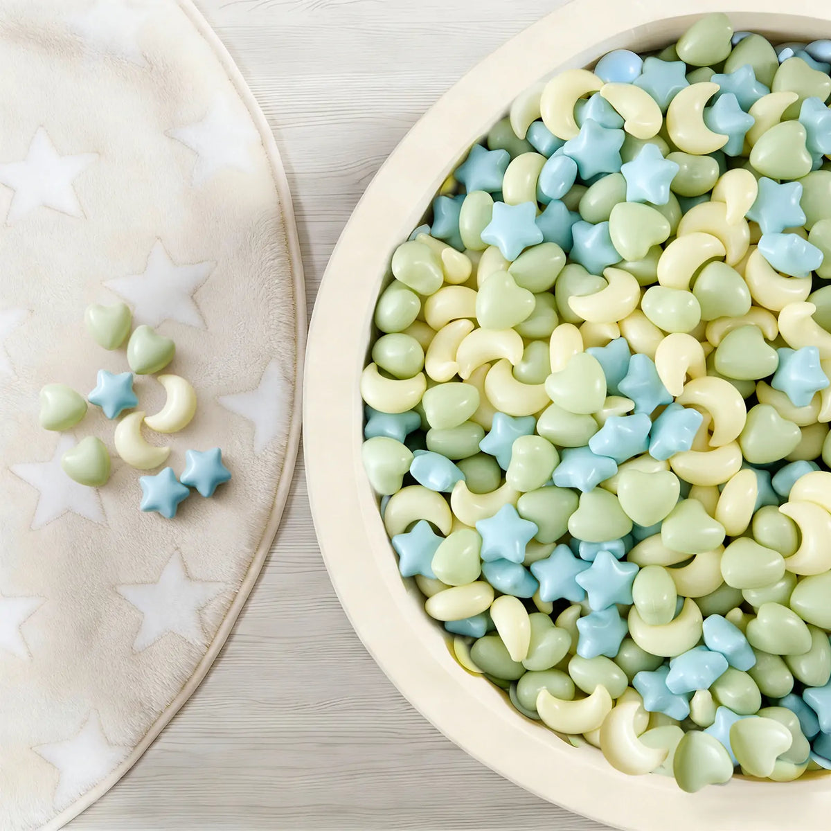 A round container filled with 200 Fun-Shapes Ball Pit Balls in light green, blue, and yellow, shaped like stars, hearts, and moons, with a few more scattered on a nearby star-patterned mat.