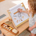 A child uses a 3-in-1 Adjustable Tabletop Art Easel to arrange wooden letters spelling SMILE below a drawing of a cactus, with a tray of supplies including numbers, markers, and an eraser nearby.