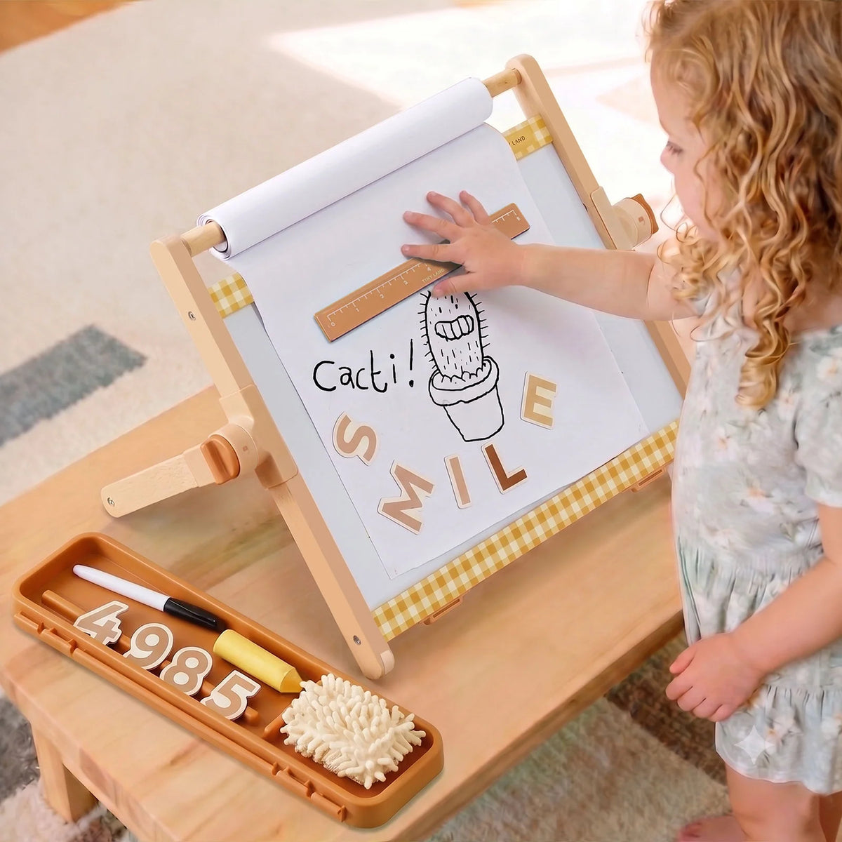A child uses a 3-in-1 Adjustable Tabletop Art Easel to arrange wooden letters spelling SMILE below a drawing of a cactus, with a tray of supplies including numbers, markers, and an eraser nearby.