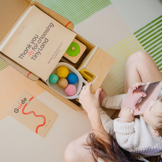 A person and a baby sit on a play mat with an open box of Tiny Land® Montessori Toys for Newborns (7–9 Months), showing colorful sensory balls, a thank-you card, a guide booklet, and the baby holding a card with a baby’s photo.