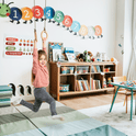 A young girl in a playroom swings on the 3in1 Swing & Climbing Set—featuring gym rings—above soft mats, engaging in activities that support child development. Behind her are bookshelves with toys and books, colorful number circles on the wall, and a table with beads.