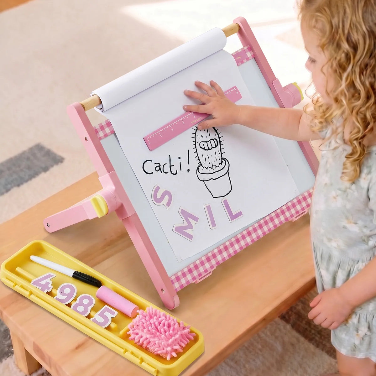 A child plays with a pink 3-in-1 Adjustable Tabletop Art Easel featuring a paper roll, a drawing of a cactus, and magnetic letters on a whiteboard, with a yellow tray containing markers and numbers nearby.