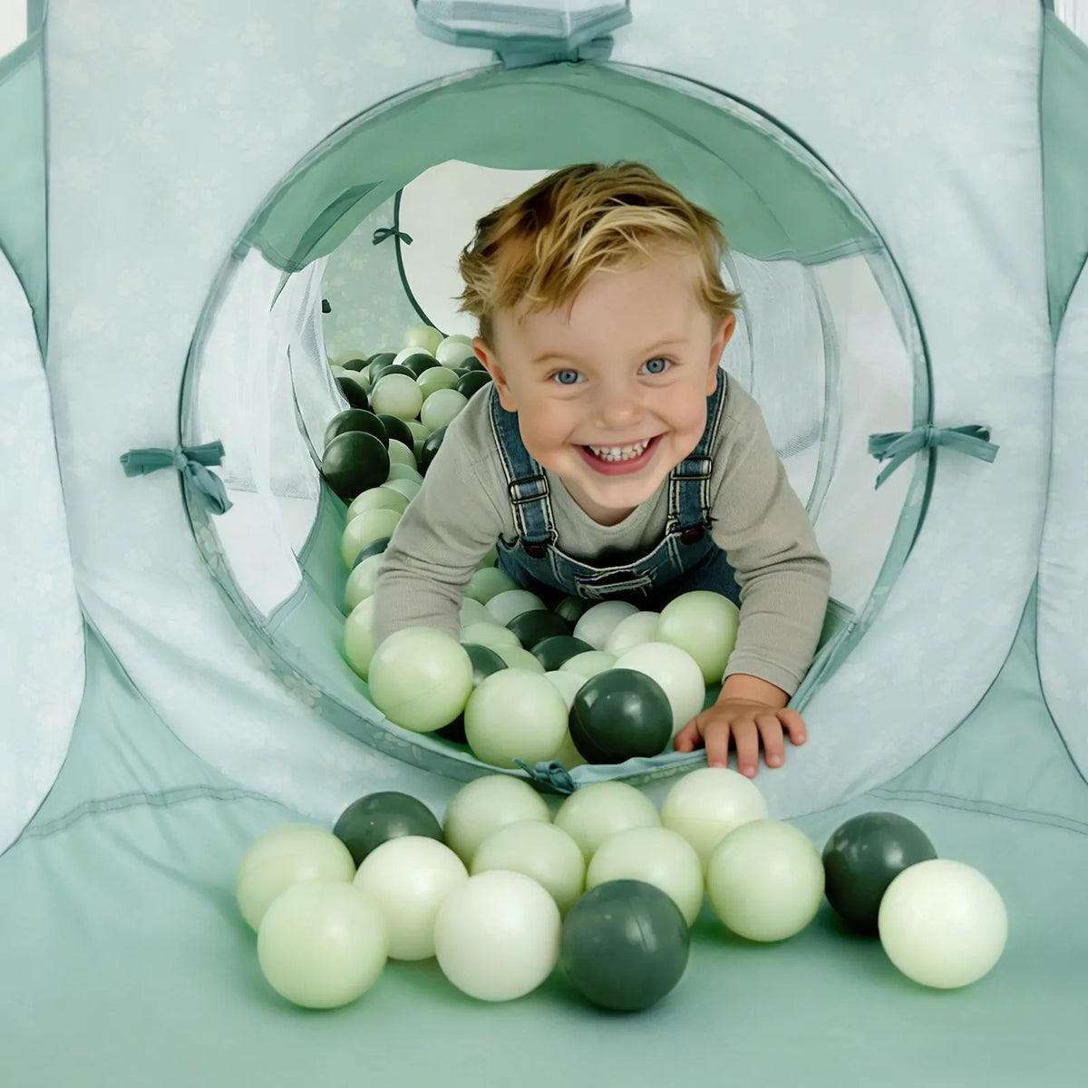 A smiling young child crawls through a fabric tunnel filled with light and dark green Ball Pit Balls (200 pc) – Soft Mixed Tones.