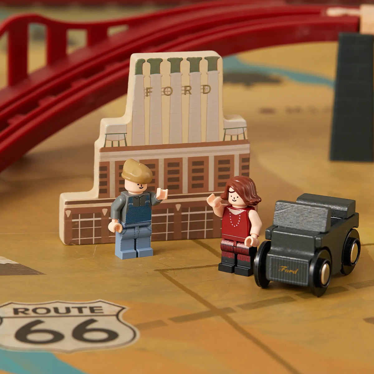 Two minifigures stand on a 1920s Railway Wooden Train Table featuring a Route 66 sign, a small black vintage car, and a factory building labeled Ford, evoking a vintage city scene.