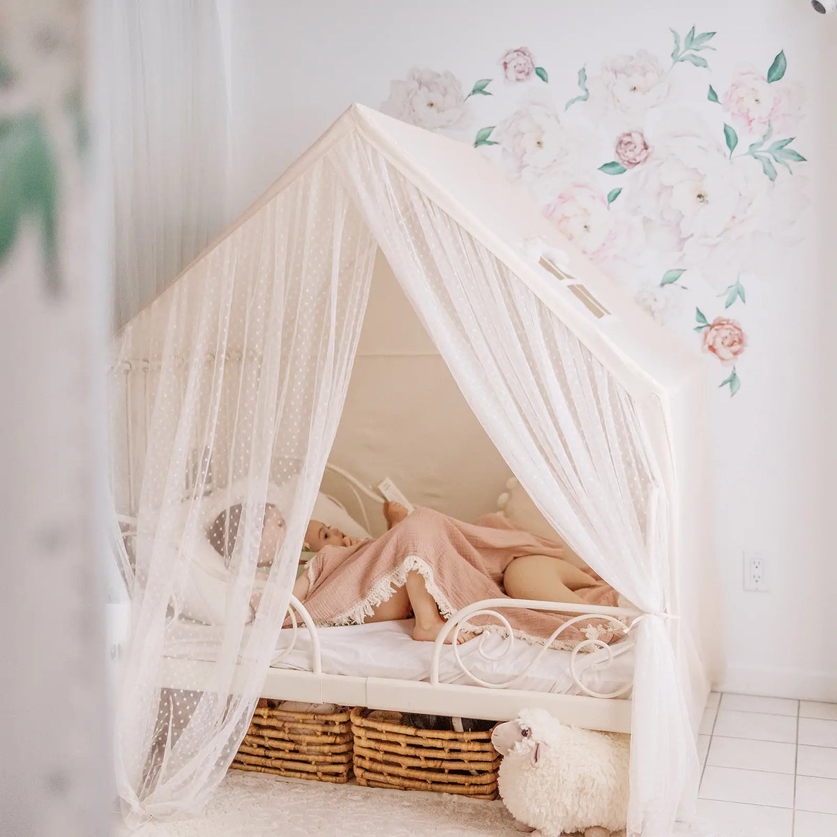 A child lies under a blanket inside a Large Cozy Cabin Play Tent with Star Lights, which is shaped like a house and draped with sheer, star-patterned curtains.