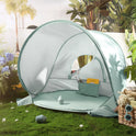 A dome-shaped Kids Beach Tent & Sand Toy Set with a green floor and light green canopy, set up on grass with a small sand toy set inside and a basket of accessories nearby.