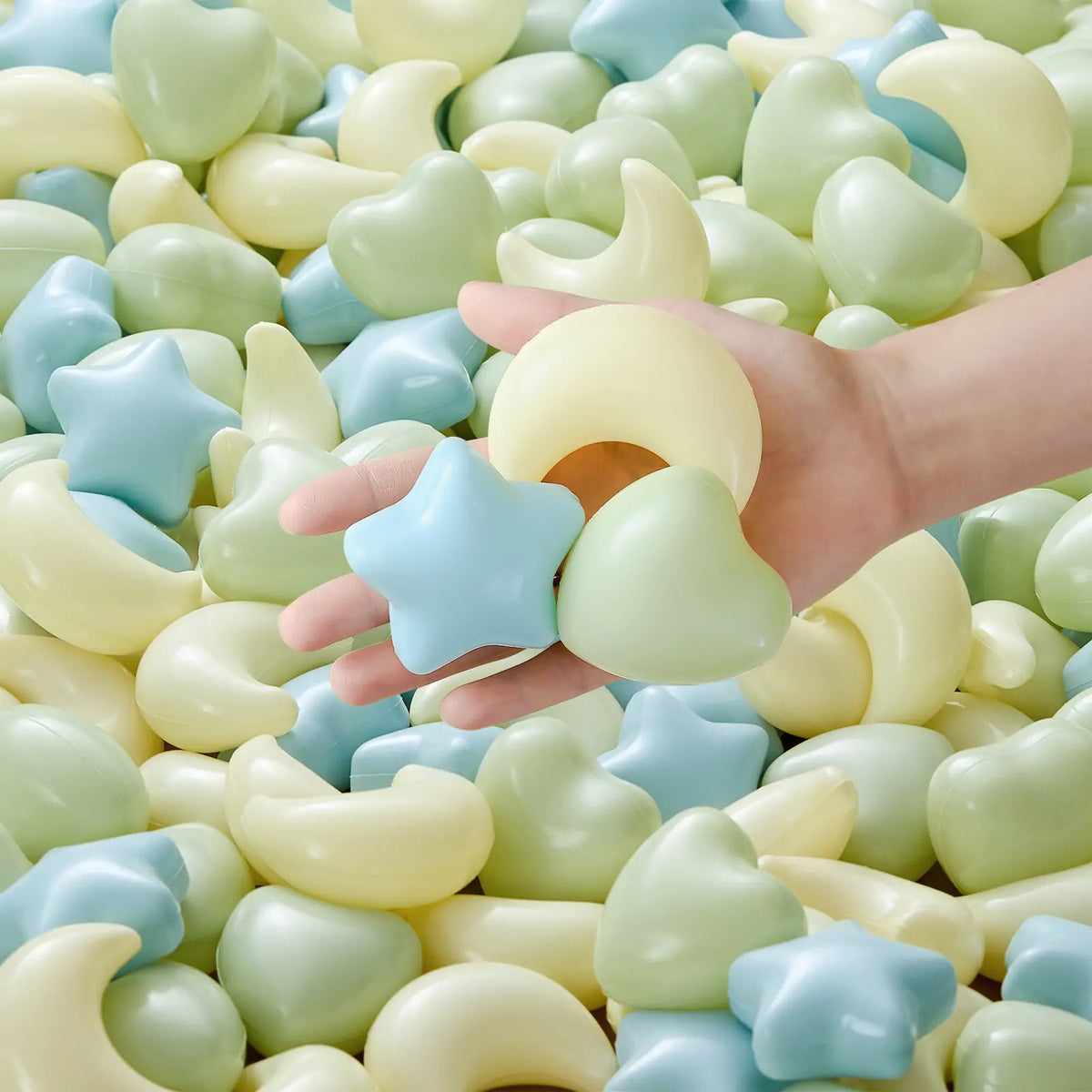 A hand holds a yellow crescent moon, a blue star, and a green heart from a set of Fun-Shapes Ball Pit Balls (200 pc), with more sensory exploration balls in the background.