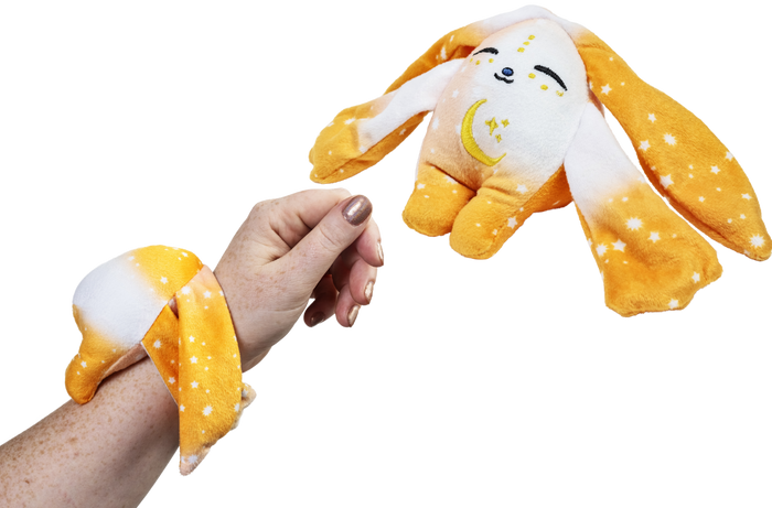 A person wears an orange, star-patterned Ori Mini Wrist Hugger – Magnetic Sensory Plush, featuring a plush wrist cuff and a matching magnetic rabbit-shaped comfort companion.