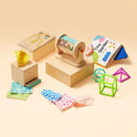 The Tiny Land® Montessori Toys for Newborns (4–6 Months) set features wooden boxes, textured fabric squares, a rolling drum toy, soft cloth book, and plastic geometric shapes in green, blue, and pink on a beige surface.