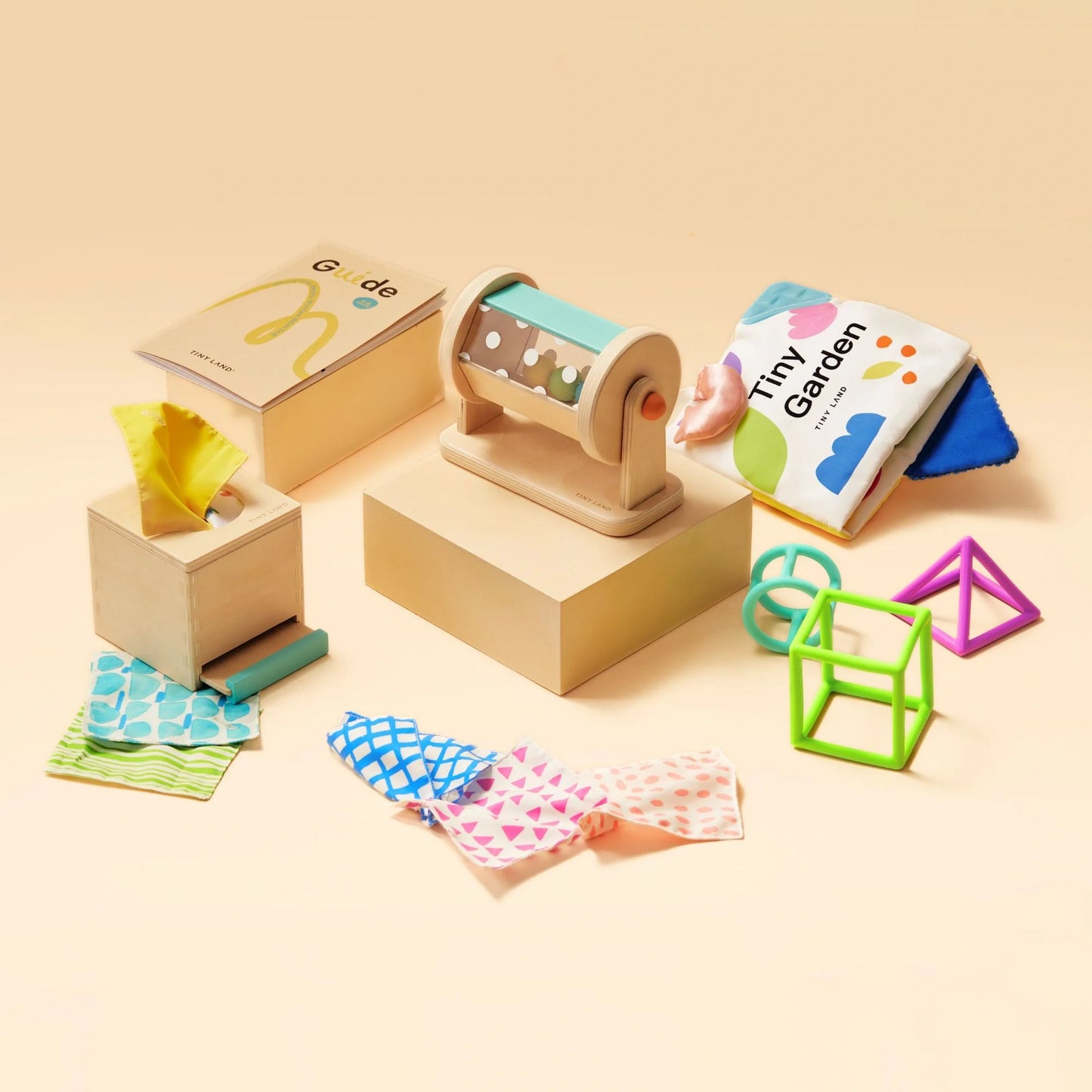 The Tiny Land® Montessori Toys for Newborns (4–6 Months) set features wooden boxes, textured fabric squares, a rolling drum toy, soft cloth book, and plastic geometric shapes in green, blue, and pink on a beige surface.