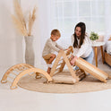 A young boy climbs the beige 4in1 Montessori Climbing Set (Triangle Ladder, Climbing Arch, Slide Board & Cushion for ages 1-3) while a woman supports him in a cozy, sunlit room with pampas grass and a woven rug.