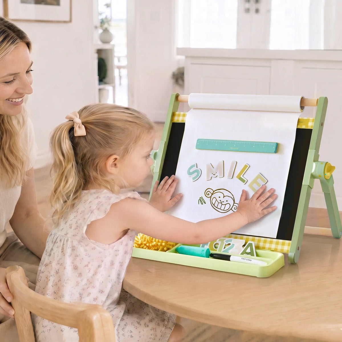 A young girl places magnetic letters that spell SMILE onto a 3-in-1 Adjustable Tabletop Art Easel while her mother watches.