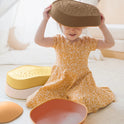 A child sits on the floor wearing a bowl-shaped Stacking Balance Stepping Stones – Sunset as a hat, surrounded by other colorful, stacking stepping stones.