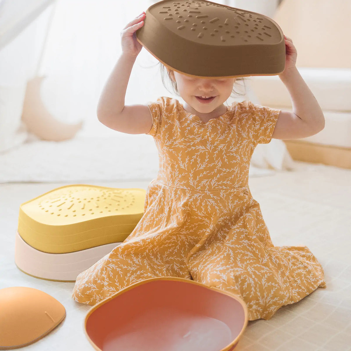 A child sits on the floor wearing a bowl-shaped Stacking Balance Stepping Stones – Sunset as a hat, surrounded by other colorful, stacking stepping stones.